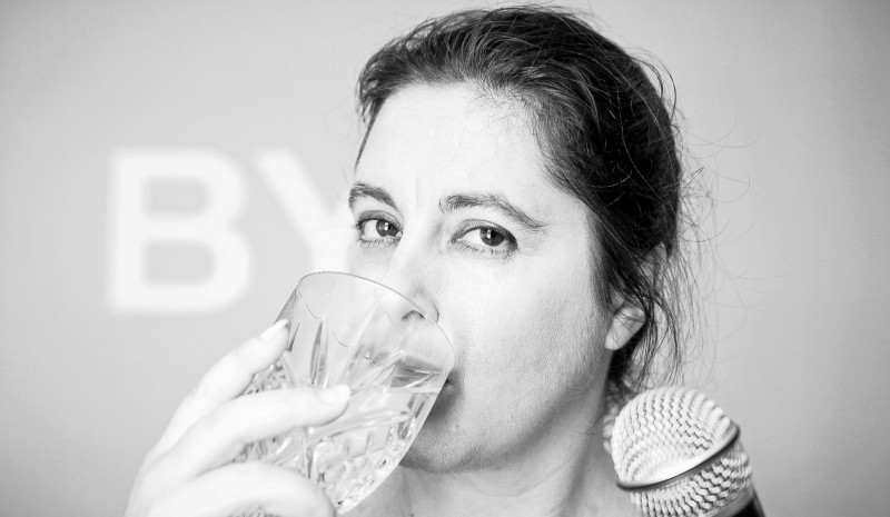 In a black and white picture, Anabella Lenzu drinks from a glass of water while holding a microphone